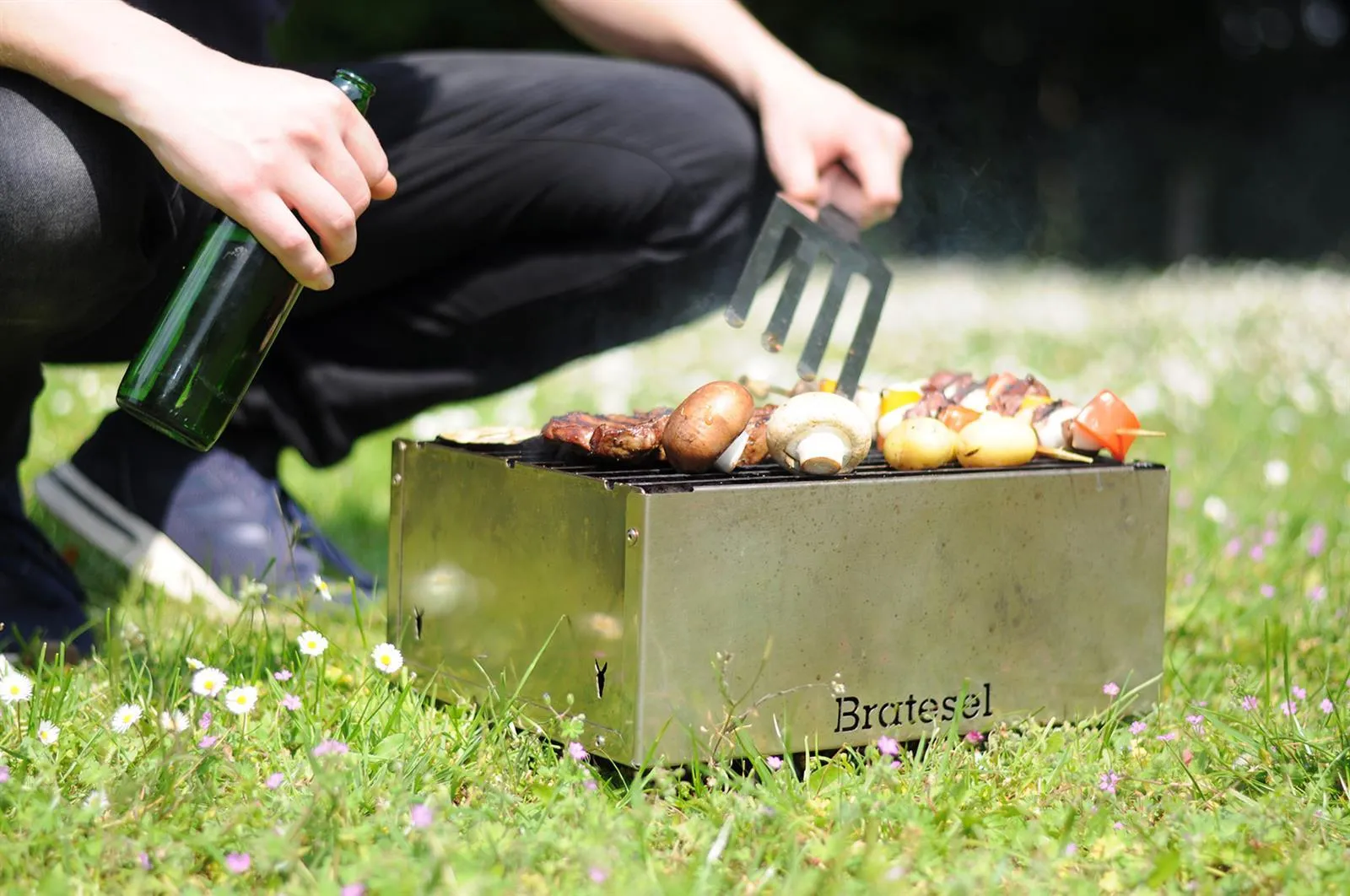 Person grillt Gemüse auf dem Bratesel Holzkohlegrill auf einer Blumenwiese